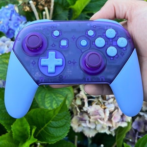 May include: A purple and light blue video game controller. The controller features a cross-shaped directional pad, two analog sticks, and various buttons. The grips and buttons are light blue, with a transparent purple body.