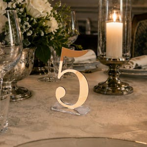 May include: A reflective rose gold table number five sign standing on a clear acrylic base, positioned on a formal dining table set with white floral arrangements, crystal wine glasses, a lit pillar candle, and white dinner plates.