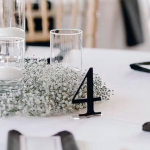 May include: A white table centrepiece with a cluster of white baby's breath flowers surrounding three clear glass candle holders with white candles. A black number four table number is placed on the table.