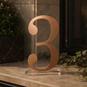 May include: A metallic rose gold number three sign with a smooth, reflective finish. The numeral is mounted on a clear, rectangular acrylic base, providing a stable foundation for the upright display of the digit on a flat surface.
