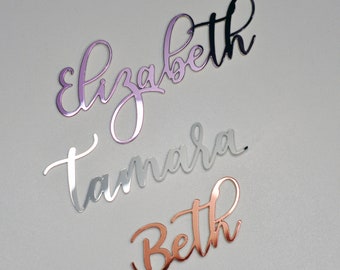 Puede incluir: Tres nombres, Elizabeth, Tamara y Beth, están escritos en cursiva sobre un fondo blanco. Los nombres están recortados en acrílico espejado y son de diferentes colores: rosa, plata y oro.