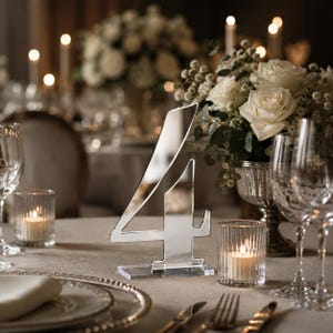 May include: A reflective, silver-toned acrylic table number four standing on a rectangular base. The number features a sharp, modern font and is positioned on a table setting with white roses, clear glassware, and ribbed glass candle holders.