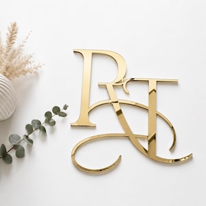 Puede incluir: Un monograma de acrílico dorado reflectante que presenta las letras mayúsculas entrelazadas R y T. El diseño muestra un acabado pulido similar a un espejo con curvas elegantes que conectan ambas letras en una sola pieza decorativa.