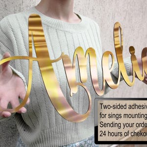 May include: A gold-coloured, reflective, script-style name sign displaying the name Amelia in a cursive font. The sign is held horizontally and features a smooth, metallic finish. A black rectangular box with white text is included in the corner.