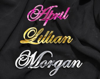 May include: Three names, "April", "Lillian", and "Morgan", are written in cursive script on a black fabric background. The names are made of mirrored acrylic, with "April" in pink, "Lillian" in gold, and "Morgan" in silver.