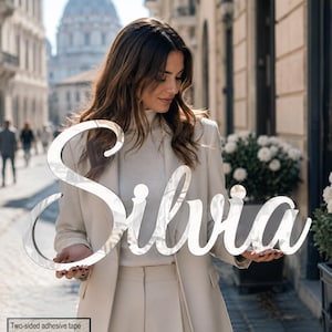 May include: A reflective, silver-toned, script-style sign featuring the name Silvia in a cursive font. The sign is held horizontally, showcasing a smooth, mirror-like surface that reflects the surrounding outdoor environment and light.