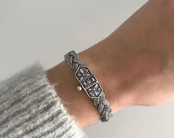 May include: A silver bracelet with a braided chain and a decorative clasp. The clasp is detailed with intricate floral patterns.