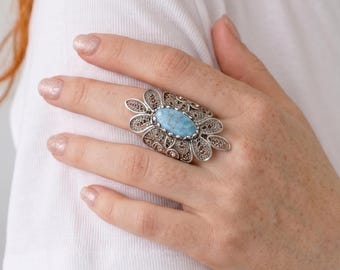 Puede incluir: Un anillo de plata con una piedra grande, ovalada y azul claro en el centro, rodeada por un diseño floral de filigrana. El anillo se lleva en una mano, con una camiseta blanca de fondo. El diseño del anillo es intrincado.
