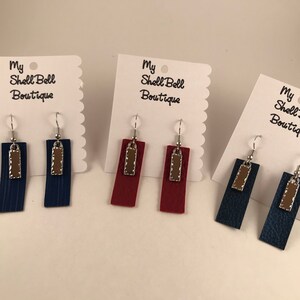 Peut inclure: Trois paires de boucles d'oreilles en cuir avec un accent en cuir marron. Les boucles d'oreilles sont présentées sur des cartes blanches avec le texte "My Shell Bell Boutique".