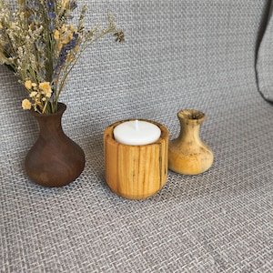 May include: A wooden candle holder with a lit white candle, a small wooden vase, and a dark brown wooden vase with dried flowers. The candle holder is cylindrical and made of light-coloured wood.