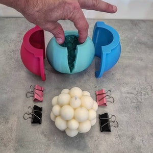 May include: A teal blue silicone mold for making a round candle. The mold is split in half and is open to show the inside. The mold is sitting on a white surface with a white candle made in the mold. The candle is made of small white balls.