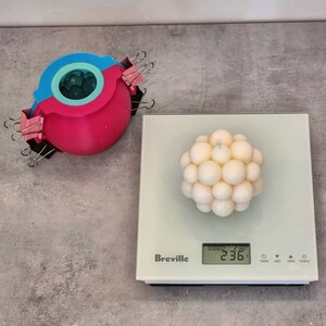 May include: A white candle shaped like a sphere with many small round bumps. The candle is on a digital kitchen scale that reads 236 grams. The scale is white with a black display and the brand name Breville.