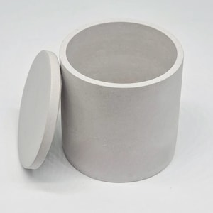 Pode incluir: Um recipiente cilíndrico de concreto cinza com uma tampa. O recipiente é feito de um material áspero e texturizado.