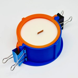 May include: A blue and orange round candle mold with a white candle inside. The mold has two metal clips attached to it.