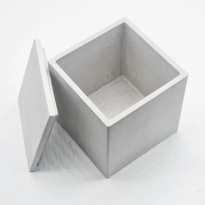 May include: A square concrete box with a lid. The box is a light grey colour and has a smooth finish. The lid is slightly ajar, revealing the empty interior of the box.