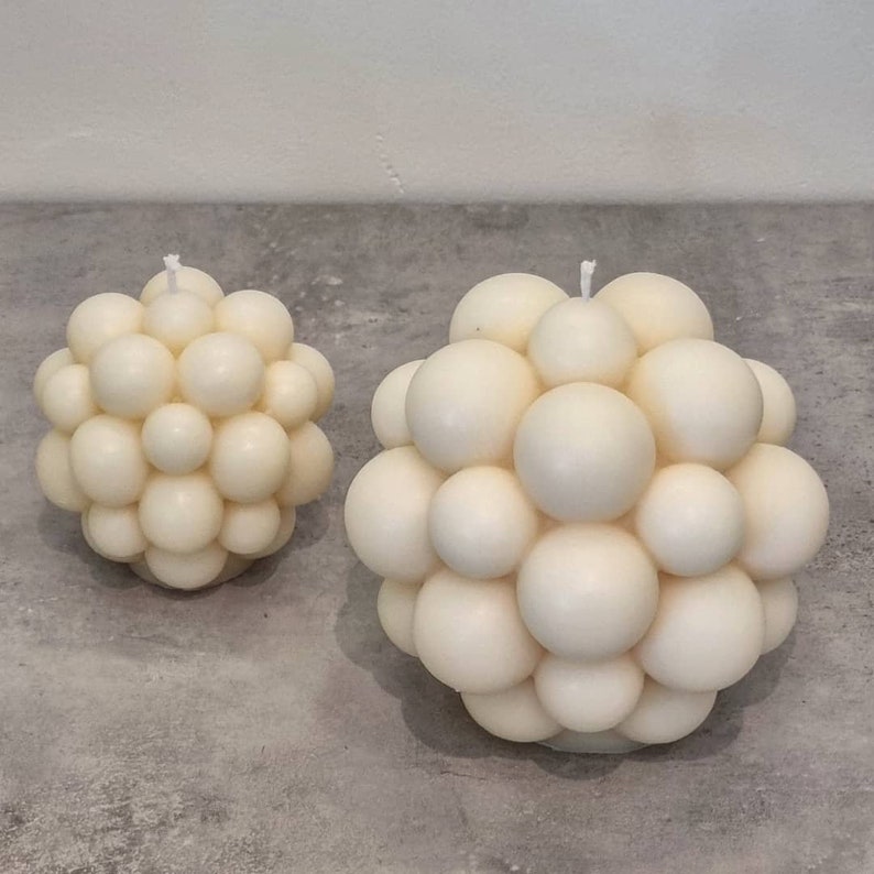 May include: Two white bubble candles, one larger than the other, on a gray surface. The candles are made of many small, round, white wax balls.