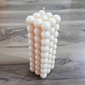 May include: A white pillar candle made of small, round balls of wax. The candle is approximately 15 cm tall and 7.5 cm wide.