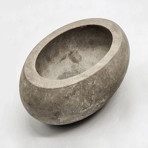 May include: An oval-shaped concrete bowl with a textured, porous gray surface. The vessel features a smooth, rounded rim and a deep, hollow interior, showcasing the natural, raw finish of the material with visible small air pockets throughout.
