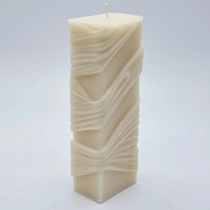 Puede incluir: Una vela blanca en forma de pilar con un diseño texturizado y en espiral. La vela es alta y rectangular.