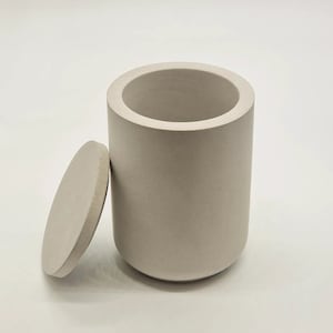 Pode incluir: Um recipiente de concreto cinza claro com uma tampa. O recipiente é cilíndrico e tem uma superfície lisa ligeiramente texturizada. A tampa é redonda e encaixa perfeitamente na parte superior do recipiente.