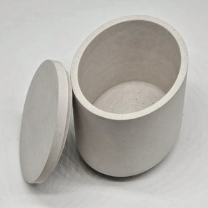 Pode incluir: Um recipiente de concreto branco oval com uma tampa. O recipiente é feito de um material áspero e texturizado.