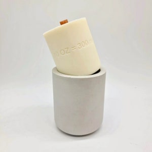 May include: A cream-colored candle with a wooden wick, labeled "10 OZ = 300 m", sits inside a gray concrete holder. The candle is cylindrical and angled, showcasing its design. The holder is a simple, rounded cylinder.