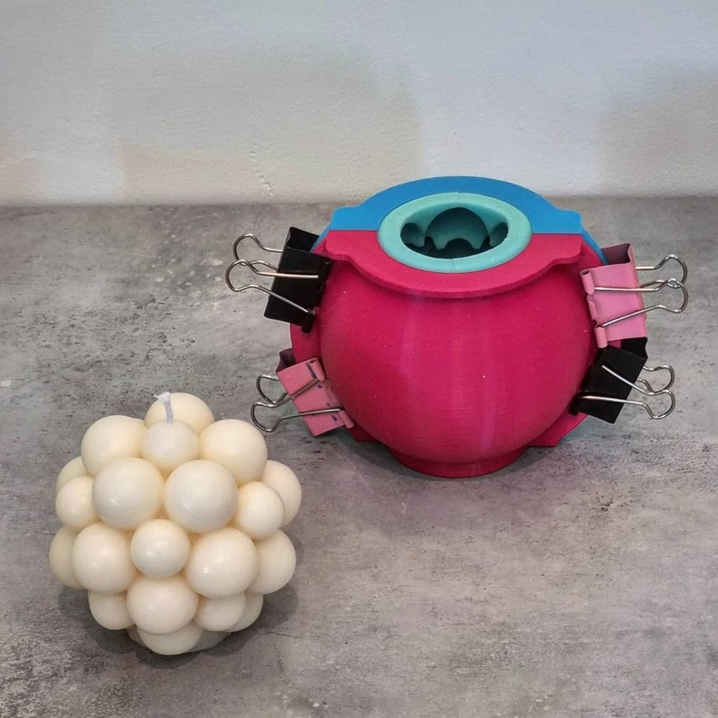 May include: A white candle with a round, bubble-like design. The candle is next to a pink and blue silicone mold with a round opening. The mold is held together with black and pink binder clips.