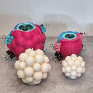 May include: Three pink and blue silicone molds for making candles. The molds are shaped like a sphere with multiple rounded bumps. Two of the molds are closed with binder clips. The third mold is filled with a white candle with a similar bumpy texture.