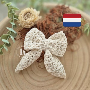 May include: A white crocheted bow with a delicate, lacy pattern. The bow is displayed on a wooden surface with a small, dried flower and a miniature Dutch flag.