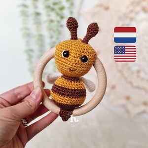 Op de afbeelding: Een gehaakte bij rammelaar met een houten ring. De bij is geel en bruin met zwarte strepen. De bij heeft een glimlachend gezicht met zwarte ogen.
