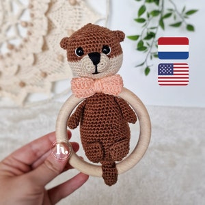 Op de afbeelding: Een bruine gehaakte otter met een roze strik die aan een houten ring is bevestigd. De otter houdt de ring vast met zijn pootjes. De ring is een babyratel.