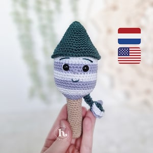 Op de afbeelding: Handgemaakte gehaakte pop met een grillig ontwerp. De pop heeft een gestreepte kop in paarse en witte tinten, een groene puntmuts en een beige steel. Het heeft zwarte ogen en een lachende mond. Een uniek, handgemaakt item.