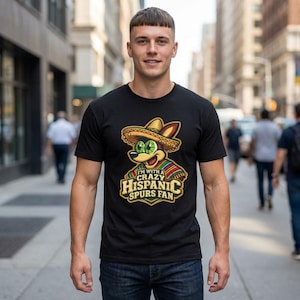 May include: Black t-shirt featuring a cartoon coyote wearing a sombrero and serape. The text on the shirt reads "I'M WITH A CRAZY HISPANIC SPURS FAN". The design is in shades of green, yellow, red, and brown.