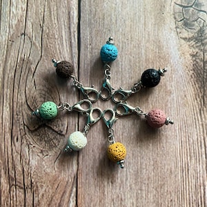 Könnte beinhalten: Sieben Lavasteine ​​in verschiedenen Farben, darunter Blau, Grün, Weiß und Braun, sind an silbernen Karabinerhaken befestigt. Die Perlen sind sternförmig auf einer Holzoberfläche angeordnet.