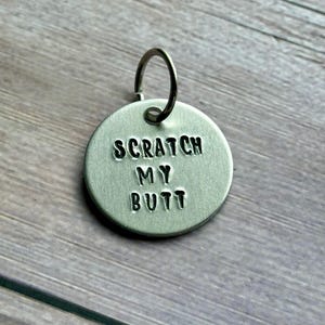 Puede incluir: Una placa redonda de color plateado para mascotas con el texto estampado "SCRATCH MY BUTT". La placa tiene un pequeño lazo de metal en la parte superior para sujetarla a un collar. El fondo es una superficie de madera.