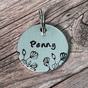 May include: A round, silver pet tag with the name "Penny" in black script. The tag features a floral design in black along the bottom edge and includes a silver ring for attachment.