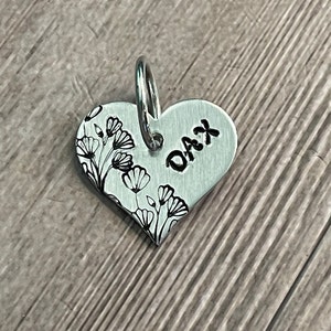 May include: A heart-shaped, silver-colored pet tag with the name "DAX" stamped on it. The tag features a floral design on the left side and a silver ring for attachment.