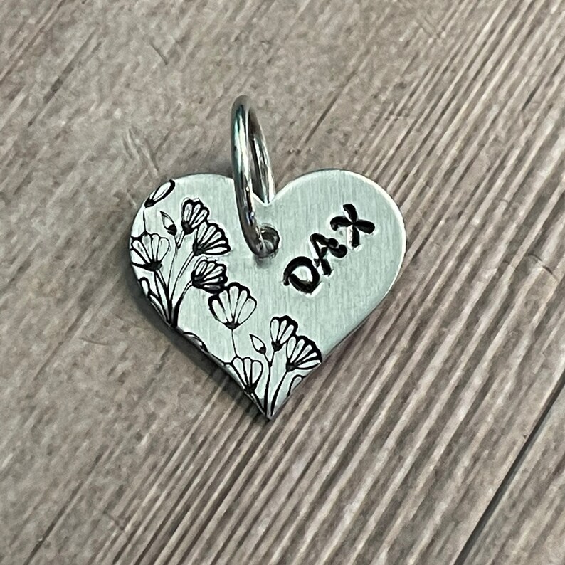 May include: A heart-shaped, silver-colored pet tag with the name "DAX" stamped on it. The tag features a floral design on the left side and a silver ring for attachment.