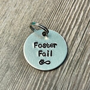 Peut inclure: Porte-clés en argent avec le texte "Foster Fail" et un symbole d'infini avec un cœur au milieu.