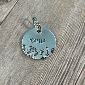 May include: A round, silver-colored pet tag with the name "Tallie" stamped on it. The tag features a floral design along the bottom edge and a silver ring for attachment.