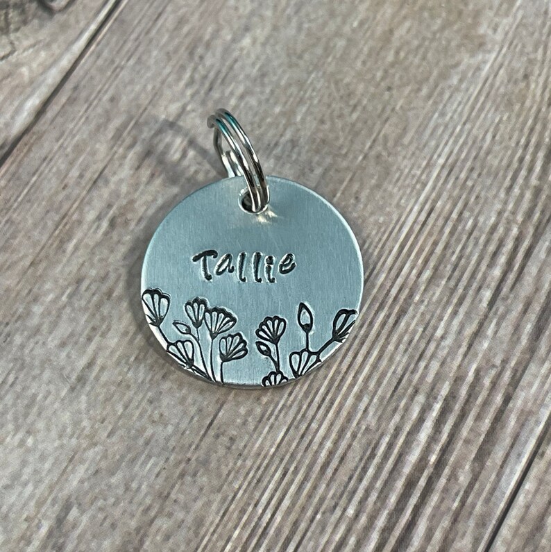 May include: A round, silver-colored pet tag with the name "Tallie" stamped on it. The tag features a floral design along the bottom edge and a silver ring for attachment.
