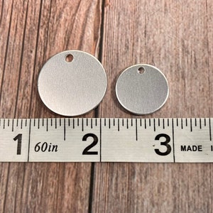 May include: Two silver-colored, round metal tags with a hole at the top, one larger than the other. A ruler shows the larger tag is approximately 2 inches in diameter. The tags are on a wooden surface.