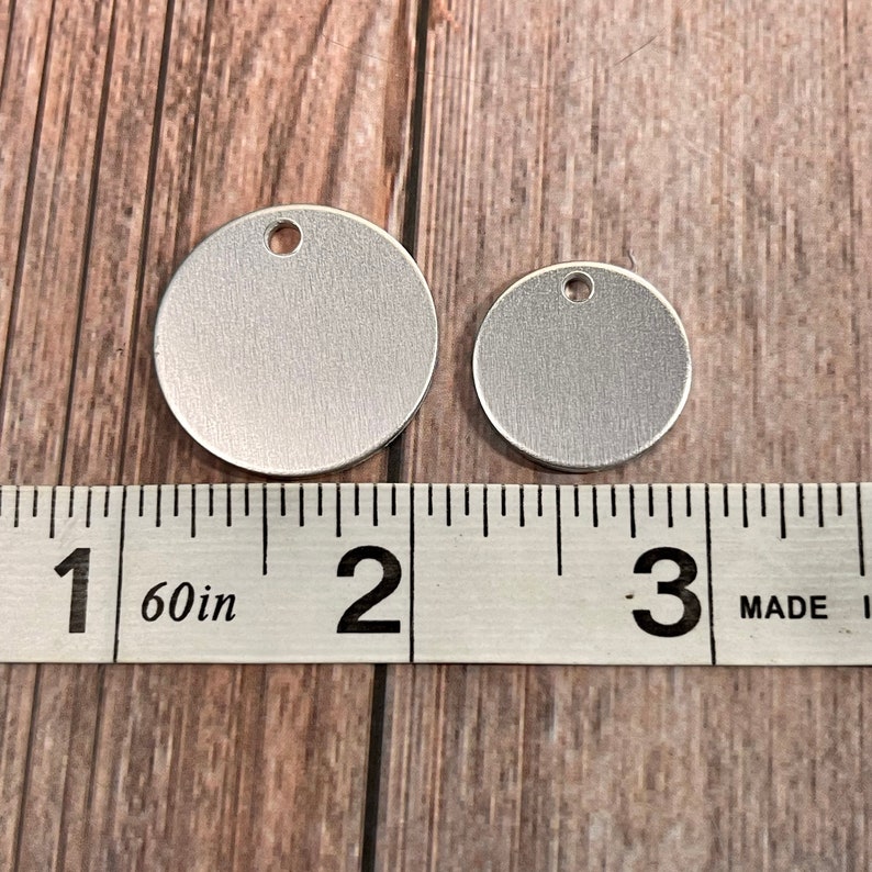 May include: Two silver-colored, round metal tags with a hole at the top, one larger than the other. A ruler shows the larger tag is approximately 2 inches in diameter. The tags are on a wooden surface.