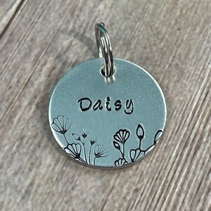May include: A round, silver-colored pet tag with the name "Daisy" stamped in the center. The tag features a floral design along the bottom edge and includes a silver ring for attachment.