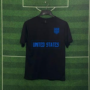 May include: Black t-shirt with blue text and a shield graphic. The shirt features the text "UNITED STATES" across the chest and "USA" above a shield logo, suggesting a sports or national theme.