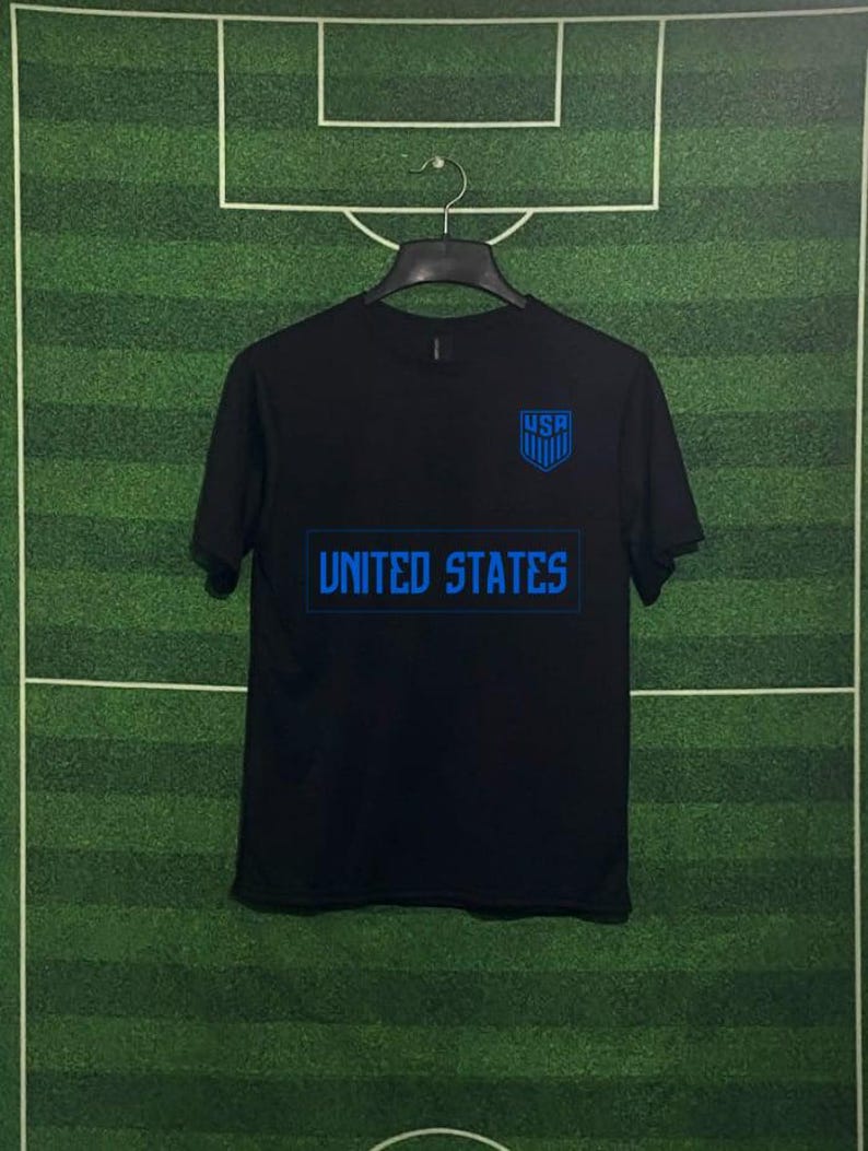 May include: Black t-shirt with blue text and a shield graphic. The shirt features the text "UNITED STATES" across the chest and "USA" above a shield logo, suggesting a sports or national theme.