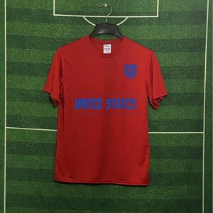 May include: A red t-shirt with the words "UNITED STATES" in blue block letters across the front. Above the text is a blue shield with the letters "USA". The shirt is hanging on a black hanger against a green background.