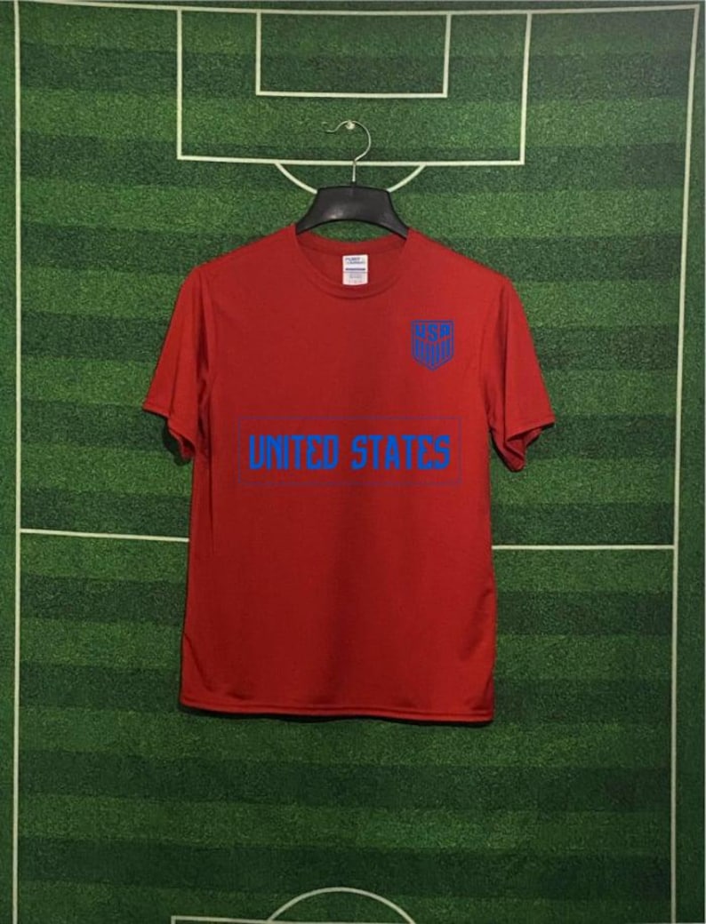 May include: A red t-shirt with the words "UNITED STATES" in blue block letters across the front. Above the text is a blue shield with the letters "USA". The shirt is hanging on a black hanger against a green background.