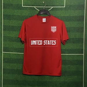 May include: A red t-shirt with the text "UNITED STATES" in a white rectangular box across the chest. Above the text is a white and blue shield with "USA" printed on it. The shirt is hanging on a black hanger against a green background.