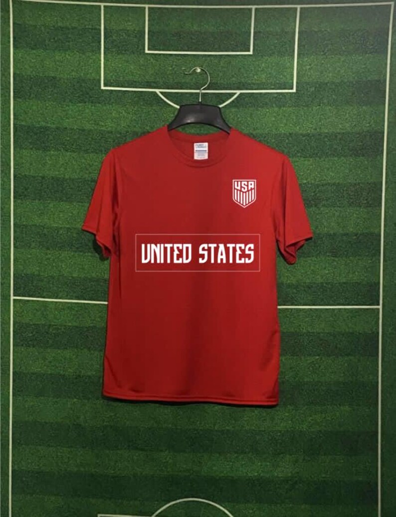 May include: A red t-shirt with the text "UNITED STATES" in a white rectangular box across the chest. Above the text is a white and blue shield with "USA" printed on it. The shirt is hanging on a black hanger against a green background.
